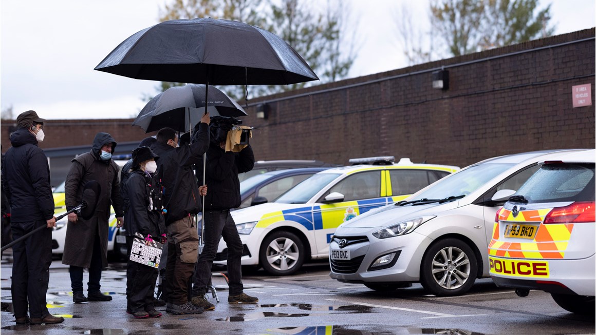 Behind the scenes production image from the high-end TV series The Responder. In the background, a row of parked police cars are seen. In the foreground, a group of production crew, all wearing dark coats and face masks, are discussing a shot, huddled under two big black umbrellas.