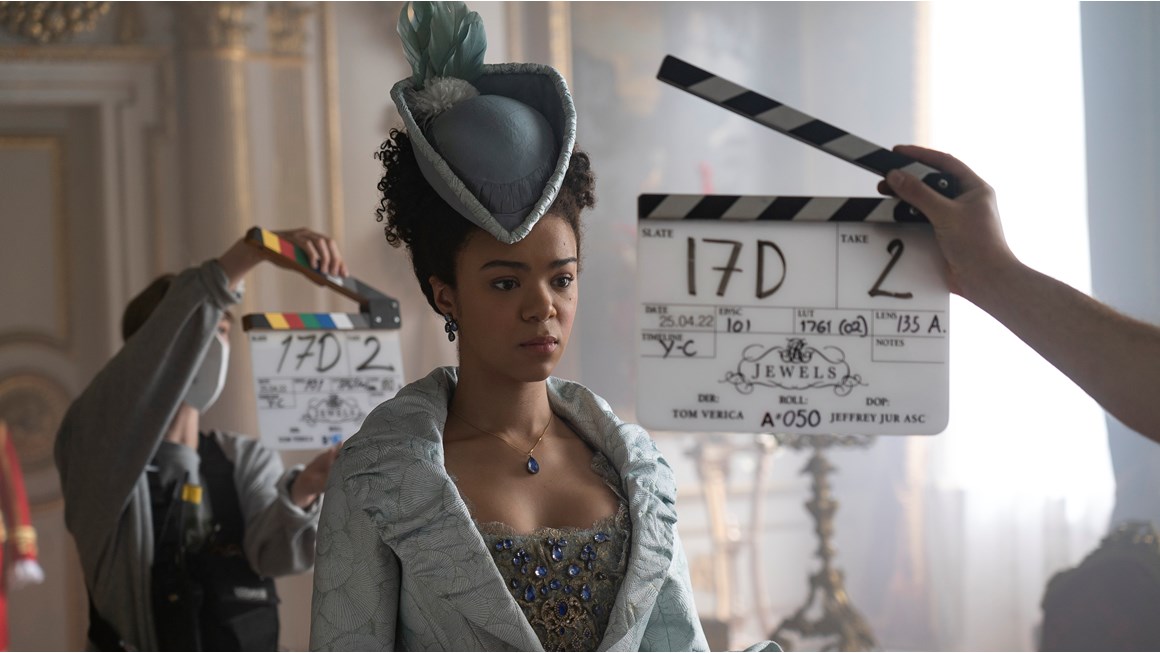 ndia Amarteifio as Young Queen Charlotte in episode 101 of Queen Charlotte: A Bridgerton Story. She is dressed in an ornate period costume in light grey with a beaded bodice and a feathered hat. Two crew members are in shot holdng clapperboards. 