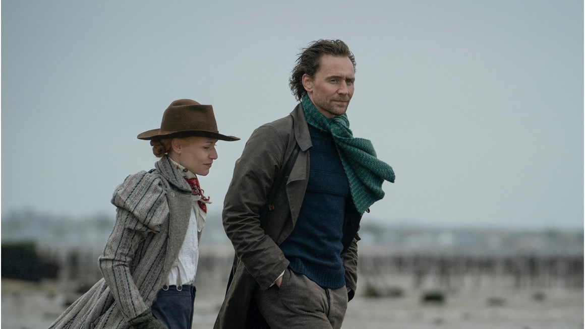 Imagae of two actors, one male and one female dressed in period costume. The Woman wears a hat, a jacket ad trusers held up by braces. The man wears a scarf and a long coat. The pair are walking outside in what appears to be a coastal location