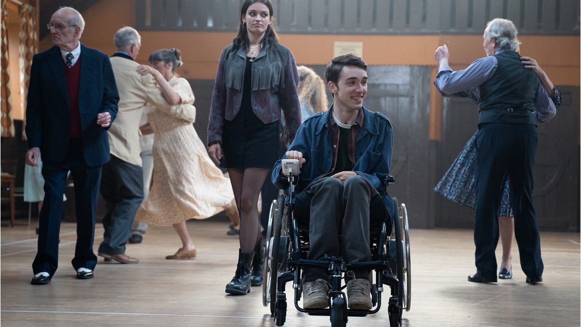 Still from Netflix TV series Sex Education featuring actors George Robinson (in wheelchair) and Emma Mackey on a dancefloor with supprting actors balroom dancing in the background