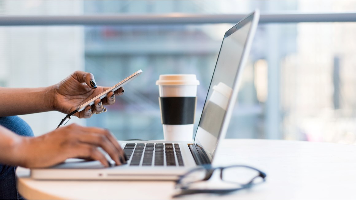 Close up of someone typing on a laptop keyboard while holding a mobile phone